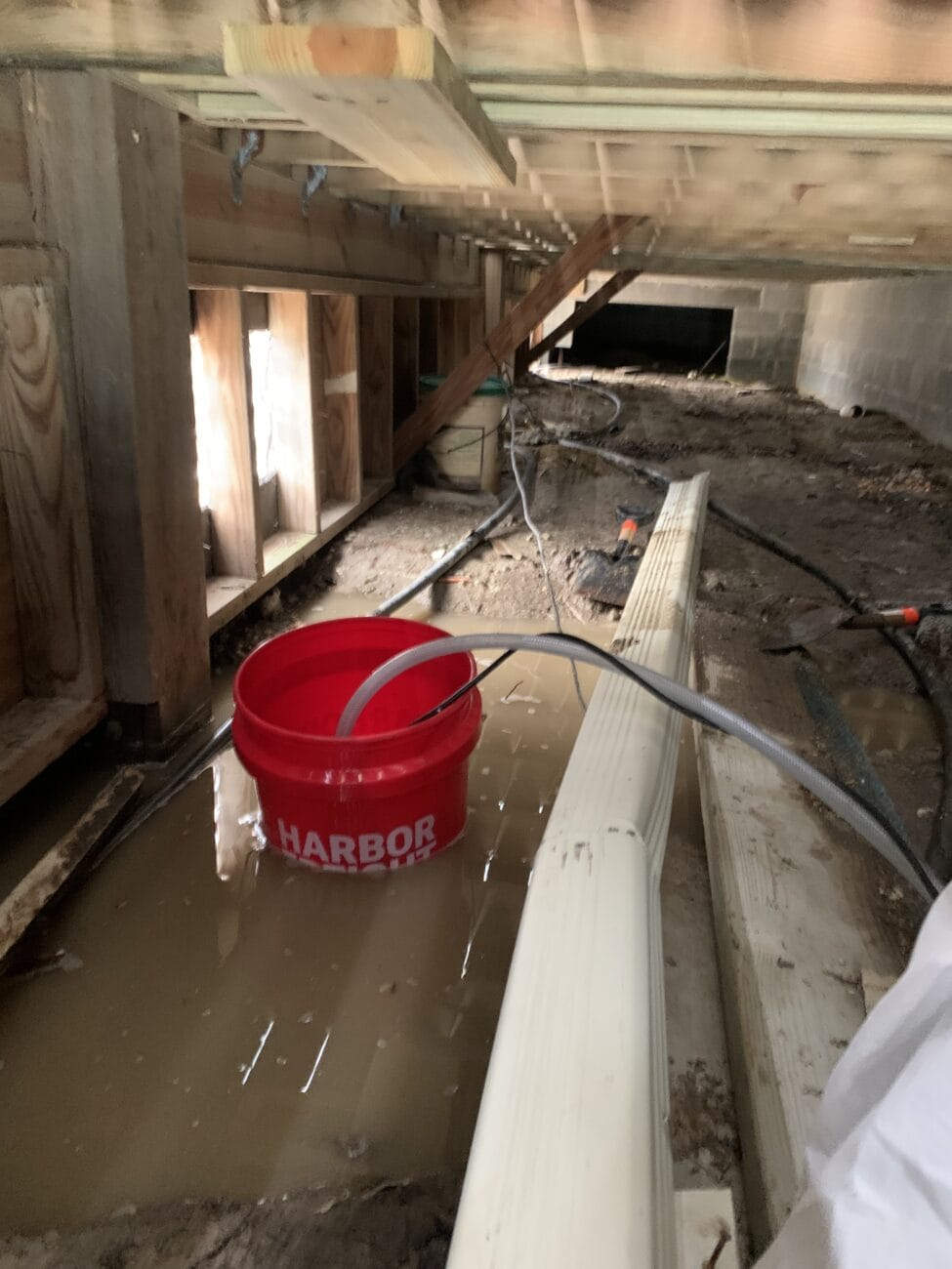 A red bucket collecting water in a muddy, flooded crawl space under a building, with pipes and construction materials scattered around.