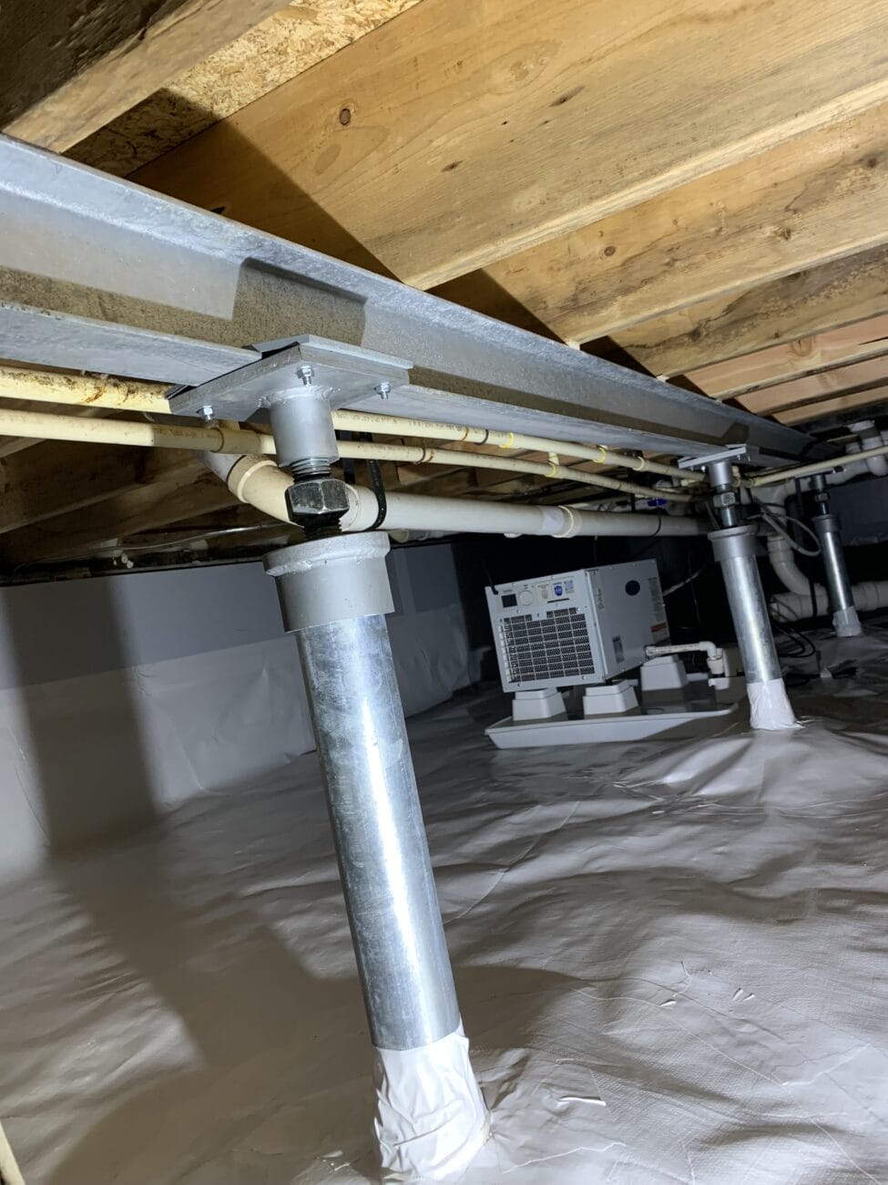 Metal support posts and pipes beneath a wooden floor, with a dehumidifier unit on a plastic-covered ground in a crawl space—ideal conditions for effective joist repair and sagging floor repair.